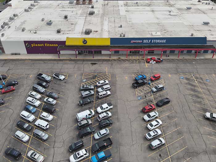 Aerial drone photograph of a commercial shopping center parking lot showing asphalt pavement, parking spaces, and vehicle traffic.