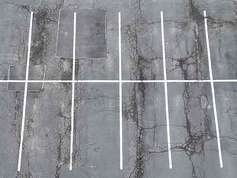 Overhead drone image showing visible asphalt cracking and pavement deterioration in a commercial parking lot.