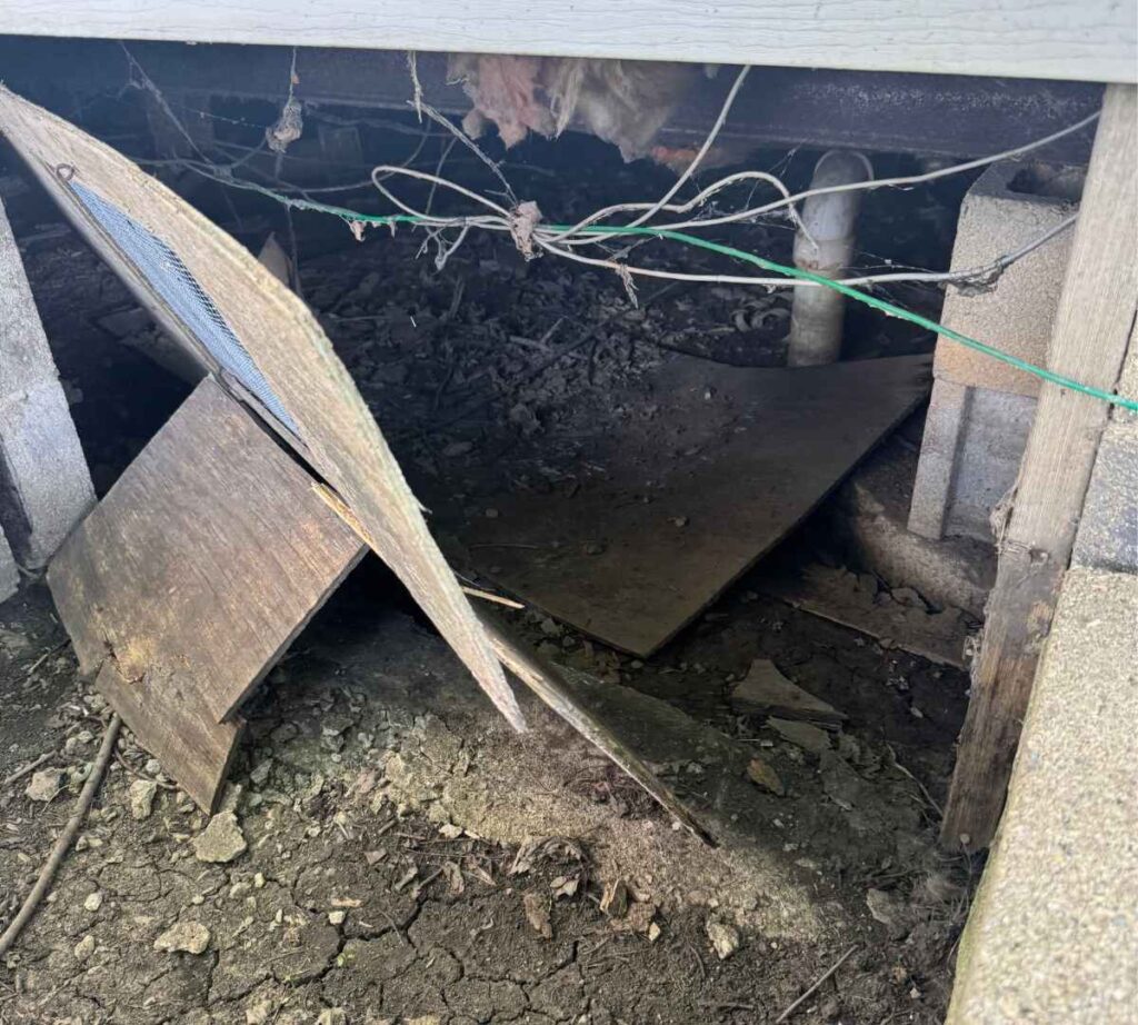 Residential crawl space with exposed soil, missing ground vapor barrier, scattered wood debris, and incomplete floor covering beneath a home.