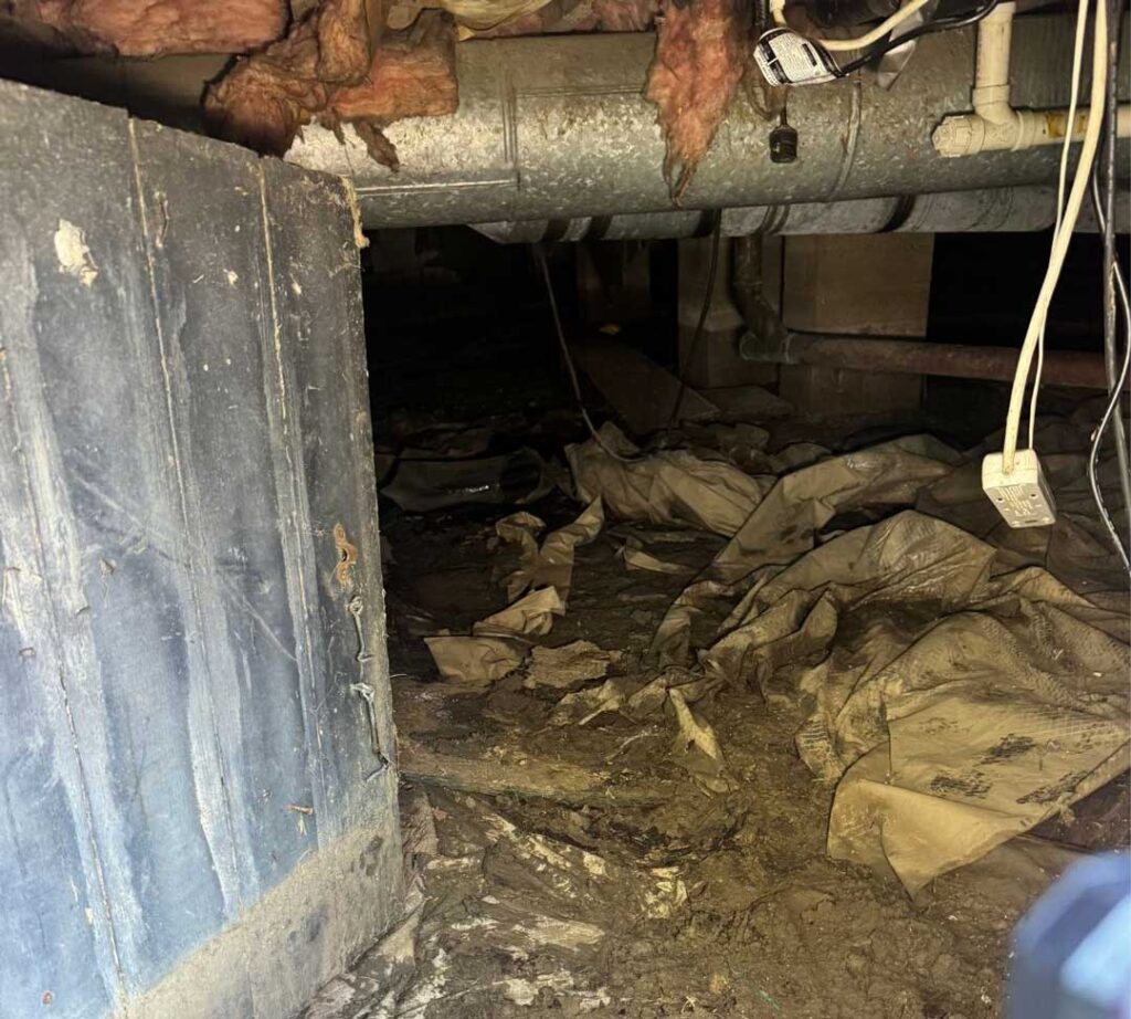 Residential crawl space with exposed soil, wrinkled ground covering, loose insulation, and overhead ductwork showing potential moisture-control concerns.