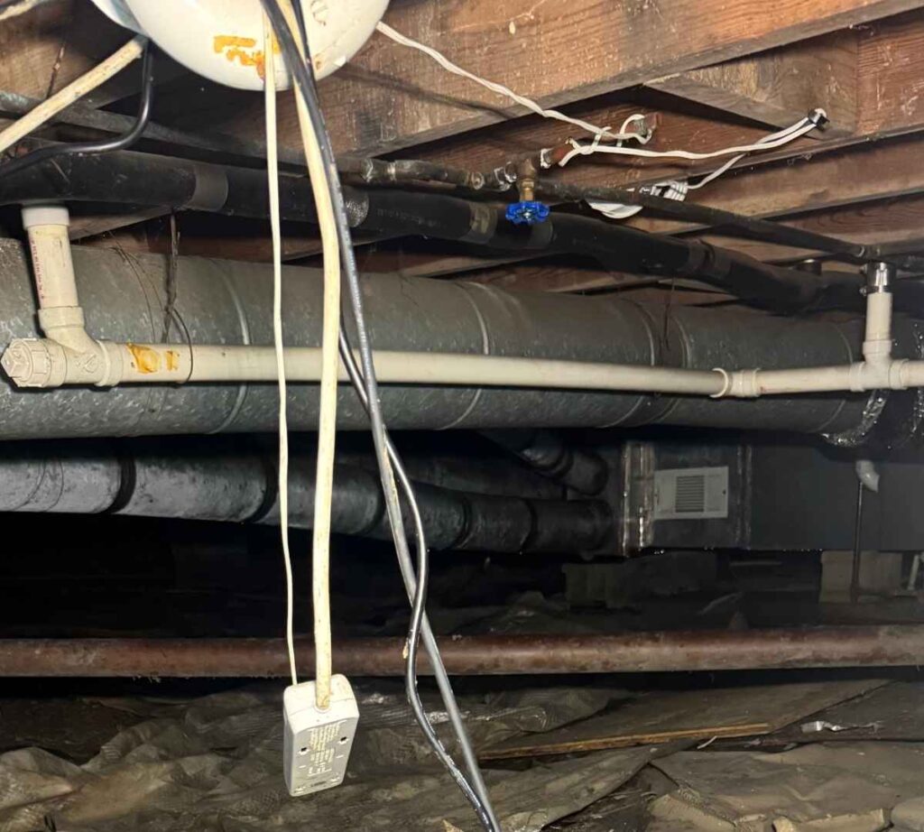 Residential crawl space showing exposed plumbing pipes, HVAC ductwork, hanging electrical wiring, and wooden floor framing beneath a home.