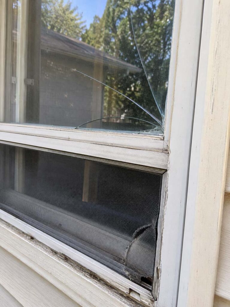 Residential window showing cracked glass, a torn insect screen, and deteriorated frame components.
