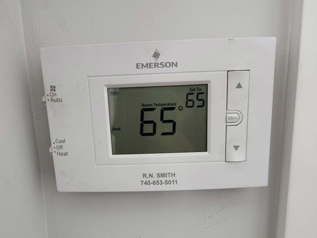 Wall-mounted Emerson digital thermostat displaying a room temperature of 65°F and thermostat settings for a residential heating and cooling system.
