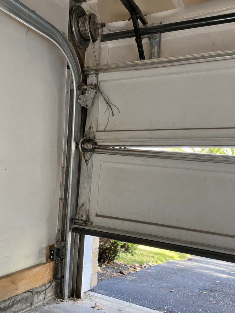 Interior view of a residential garage door showing a frayed lifting cable and worn hardware near the track and opener components.