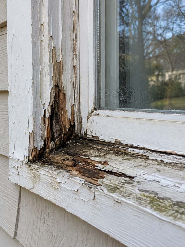 Rotted wooden window frame and sill showing peeling paint, dark moisture stains, and severe wood deterioration around a residential window.
